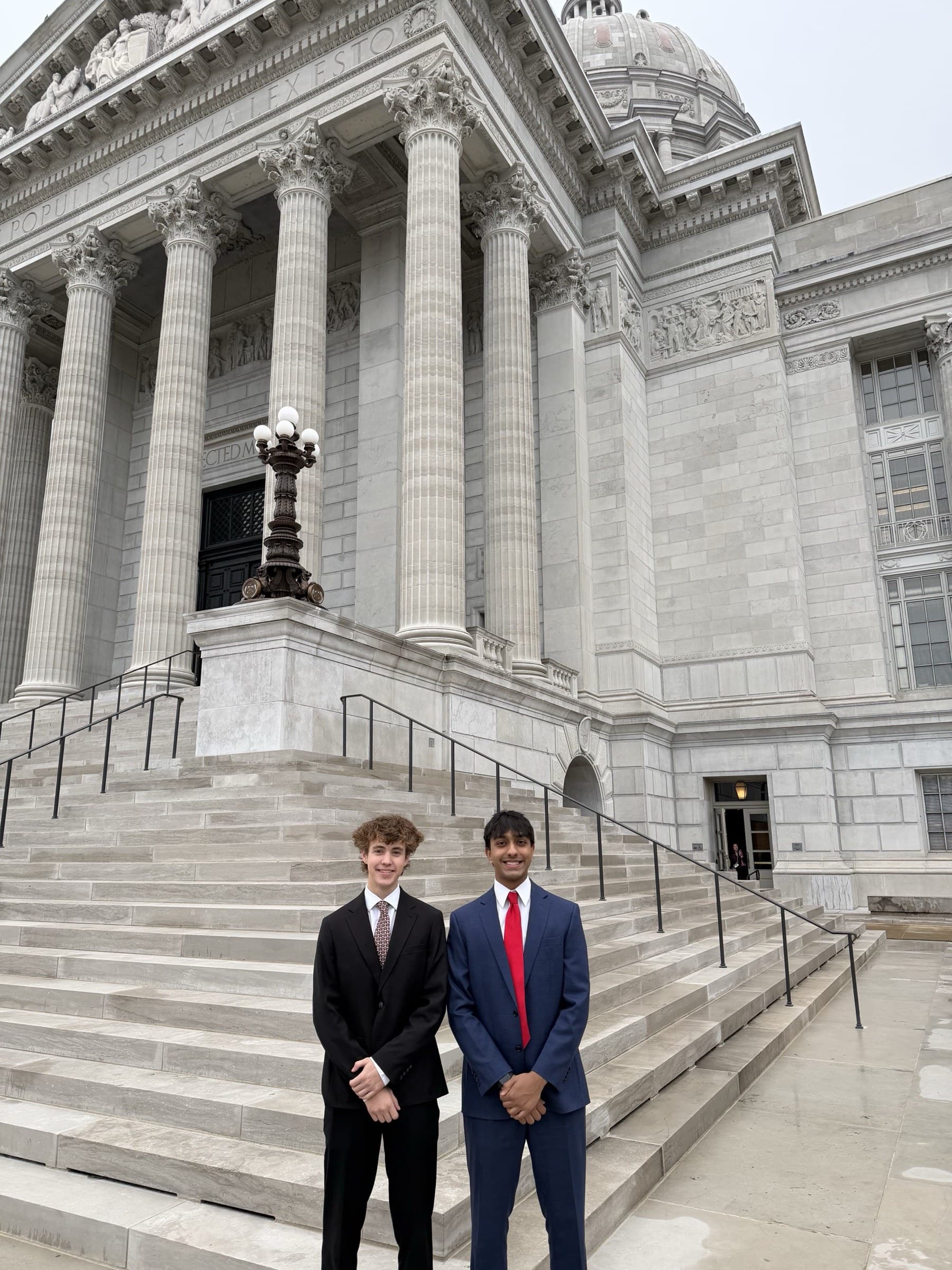 Two Atlas Health Equity Group representatives in suits standing on the steps of a state capitol building, in front of its columned entrance.